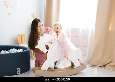 Ein kleines Mädchen von 1 Jahr in einem rosa Kleid sitzt auf einem Holzpferd, sie ist glücklich und lächelt. Ihre Mutter sitzt auf dem Boden neben ihr Stockfoto