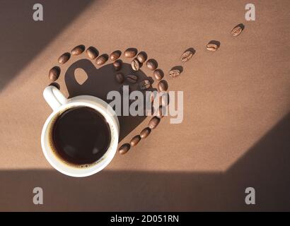 Weiße Tasse Kaffee und Kaffeebohnen mit Schatten auf braunem Hintergrund, flache Lay mit Kopierraum Stockfoto