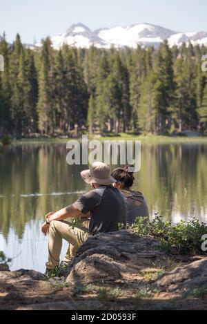 Ein junges Paar (Mann und Frau) sitzt auf Felsen neben einem See in der Morgensonne mit Bäumen, einem See und schneebedeckten Bergen in der Ferne in Calif Stockfoto
