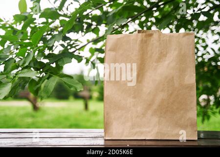 Leere Papiertüte steht auf einem Holztisch vor dem Hintergrund grüner Blätter. Einkaufskonzept. Speicherplatz kopieren. Stockfoto