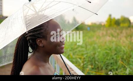 Positive afroamerikanische Frau mit großen transparenten Regenschirm steht auf leer Holzbrücke gegen grünes Schilf aus der Nähe Stockfoto