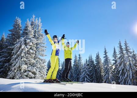 Lächelnde Frau und Mann auf Skiern mit Skistöcken in Händen. Frohlockes Paar im Skigebiet bei sonnigem Winterwetter. Schneebedeckter bewaldeter Hang. Klarer blauer Himmel mit Kopierbereich. Schnappschüsse aus dem niedrigen Winkel. Stockfoto