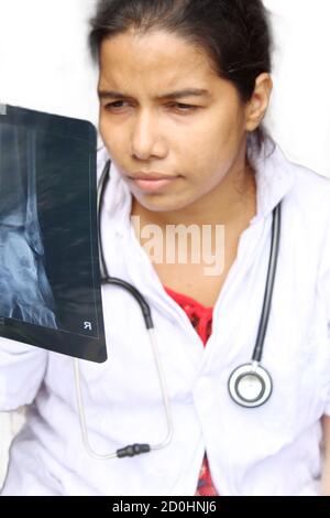 Junge Ärztin beim Betrachten von Röntgenstrahlen mit ernstem Ausdruck und weißem Hintergrund, selektiver Fokus auf Röntgenplatte, zugeschnittene Ansicht. Stockfoto