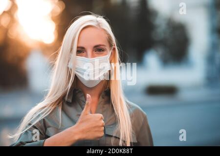 Junge blonde Frau 24-26 Jahre alt trägt medizinische Maske und zeigen Daumen nach oben Blick auf Kamera im Freien über Stadt Hintergrund. Gesundheitswesen. Coronavirus. Stockfoto