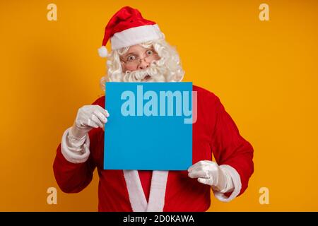 Männlicher Schauspieler in einem Kostüm des Weihnachtsmannes hält in Seine Hände ein blaues Blatt Papier für Aufzeichnungen und Posen auf gelbem Hintergrund Stockfoto