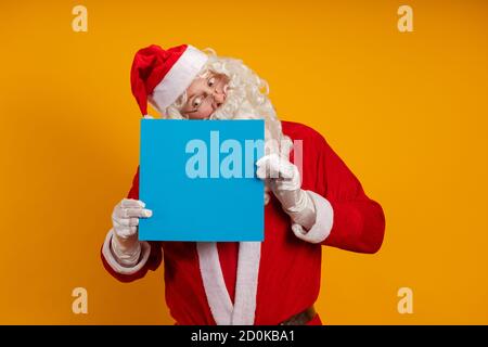 Männlicher Schauspieler in einem Kostüm des Weihnachtsmannes hält in Seine Hände ein blaues Blatt Papier für Aufzeichnungen und Posen auf gelbem Hintergrund Stockfoto