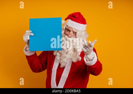 Männlicher Schauspieler in einem Kostüm des Weihnachtsmannes hält in Seine Hände ein blaues Blatt Papier für Aufzeichnungen und Posen auf gelbem Hintergrund Stockfoto