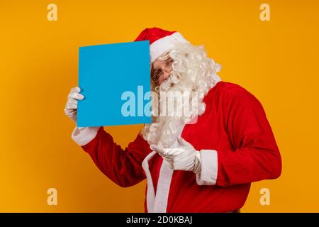 Männlicher Schauspieler in einem Kostüm des Weihnachtsmannes hält in Seine Hände ein blaues Blatt Papier für Aufzeichnungen und Posen auf gelbem Hintergrund Stockfoto