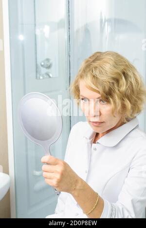 Frau mittleren Alters in weißem Medizinmantel Blick in den Spiegel. Arzt weiblich, Ärztin Stockfoto
