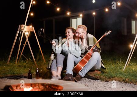 Glückliches kaukasisches Paar, Brünette Frau und bärtiger Mann mit Gitarre bei Picknick auf dem Land. Lustiges Paar macht Selfie, lächelt und hat Spaß zusammen Stockfoto