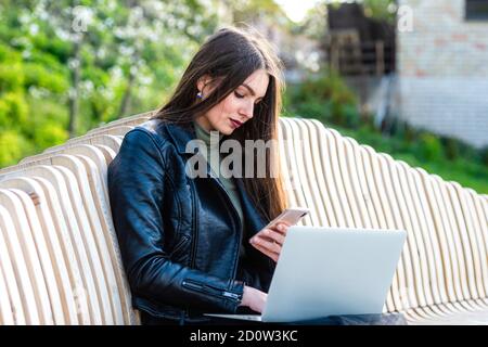 Junge elegante Frau auf Bank im Park sitzen und arbeiten auf Laptop an einem sonnigen und windigen Tag, das Konzept der Arbeit außerhalb des Büros Stockfoto