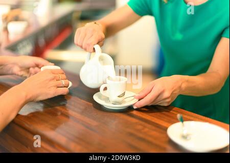 Frau hält einen Milchkännchen und eine Tasse Kaffee, gießt Schlagmilch in Espresso - Lifestyle at Coffee Shop Konzept - Fokus auf Hände Stockfoto