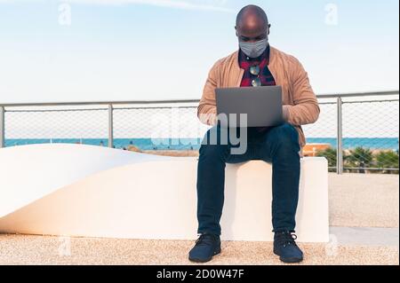 Schwarzer Mann mit der Gesichtsmaske, Arbeit auf Laptop sitzen auf einer Bank im Park - afroamerikanischen Geschäftsmann Blick auf Laptop-Bildschirm Gespräch mit Fr. Stockfoto