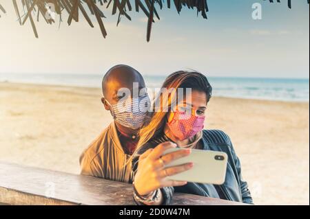 Multirassische Millennial Freunde unter Selfie lächelnd hinter Gesichtsmasken an Bar am Strand - Frohe Freundschaft und neue Normalität Konzept mit jungen Menschen Stockfoto