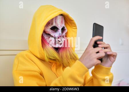 Halloween-Tag, Mädchen in gruseligen Gesichtsmaske mit Kapuze macht Videoanruf auf Smartphone Stockfoto