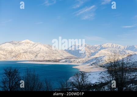 Charvak-Stausee mit blauem Wasser an einem klaren Wintertag in Usbekistan, umgeben von schneebedeckten Berggipfeln des Tien Shan Stockfoto