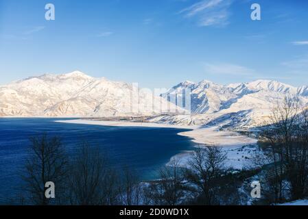 Charvak-Stausee mit blauem Wasser an einem klaren Wintertag in Usbekistan, umgeben von schneebedeckten Berggipfeln des Tien Shan Stockfoto