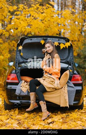 Eine junge Frau im Auto auf dem Kofferraum vor dem Hintergrund des Herbstwaldes sitzt entspannt und umarmt ihren Lieblingshund. Stockfoto