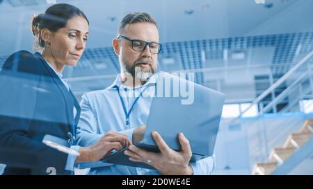 Stilvolle selbstbewusste Geschäftsfrau und Geschäftsmann arbeiten an einem Projekt, Mann hält Laptop-Computer, sie Referenz. Im Hintergrund modern und Stockfoto