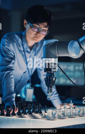 Vertikale Aufnahme eines professionellen japanischen Entwicklungsingenieurs, der eine testing Künstliche Intelligenz Schnittstelle durch das Spielen von Schach mit einem futuristischen Roboter Stockfoto