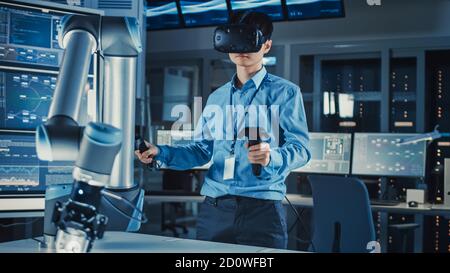 Professioneller japanischer Entwicklungsingenieur in blauem Hemd steuert ein Futuristischer Roboterarm mit Virtual Reality-Headset und Joysticks In einem Stockfoto