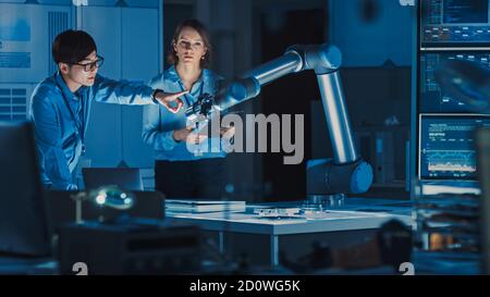 Mann und Frau Ingenieure mit Laptop und Tablet Analysieren und diskutieren, wie ein futuristischer Roboterarm funktioniert und ein Metallobjekt bewegt. Sie sind in einem hohen Stockfoto