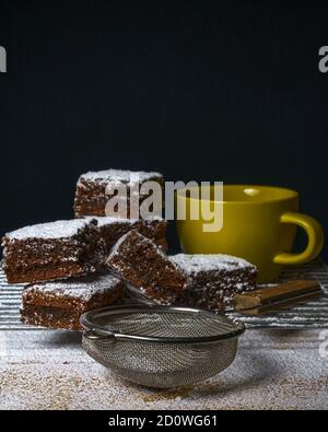 Vertikale Aufnahme von köstlichen Brownies mit Puderzucker bedeckt, neben einem Siefter und Tasse Stockfoto