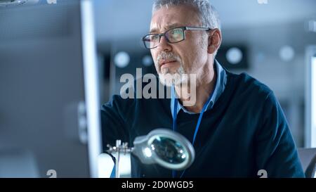 Elektronikreparaturingenieur mittleren Alters, der am Personal Computer arbeitet in seiner Werkstatt, optische Linse im Vordergrund. Im Hintergrund High Tech Stockfoto