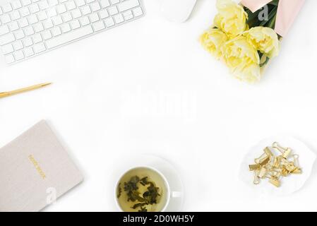 Flacher Schreibtisch für Damen. Weiblicher Arbeitsplatz mit Laptop, ein Bouquet von gelben Pfingstrosen-förmigen Tulpen, Accessoires, eine Tasse grüner Tee auf einem weißen Rücken Stockfoto