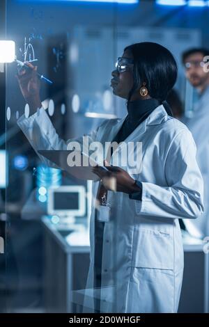 Im Forschungslabor Intelligente und schöne afroamerikanische Frau Wissenschaftler tragen weißen Mantel und Schutzbrille schreibt Formel auf Glas Stockfoto