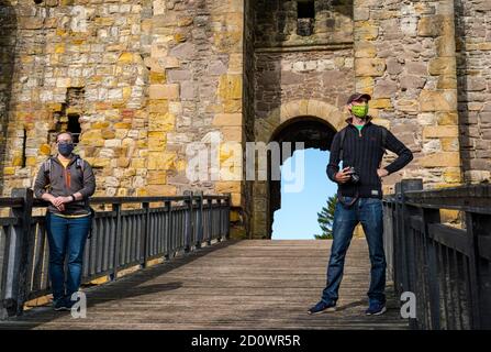 Touristen-Paar mit Gesichtsmasken stehen auf Holzbrücke über Graben am Eingang zu ruinierten mittelalterlichen Dirleton Castle, East Lothian, Schottland, Großbritannien Stockfoto