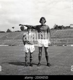 1960s, historisch, ein großer Torhüter, der mit einem anderen, kürzeren Spieler steht, der Spaß bei einem Fotoshooting vor der Saison im Valley Stadium, Southeast London, der Heimat des Charlton Athletic Footall Club, England, Großbritannien, hat. In dieser Zeit wurde Fußball auf dem Gelände mit traditionellen Terrassen gespielt, wo Fußballfans standen, um das Spiel zu beobachten, einige waren offen und einige waren überdachte Stände, wie man hier sehen kann. Stockfoto