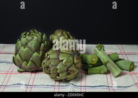 Nahaufnahme von frisch angebauten Bio-Artischocken und geschnittenen Stielen auf einem rustikalen Stück Stoff und schwarzem Hintergrund. Stockfoto