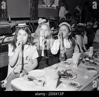 70er Jahre, historisch, drei junge Mädchen, die Spaß auf einer Kinder-Weihnachtsfeier sitzen an einem langen Tisch tragen ihre Party Hüte und bläst Luftschlangen, England, Großbritannien. Stockfoto