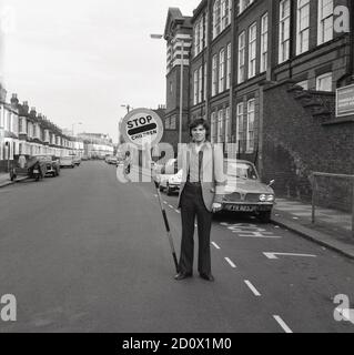 1970, historisch, ein junger Lehrer mit einem Verkehrsschild "STOP Children" Lollipop auf einer Straße vor einer großen innerLondoner Kinderschule, Southeast London, England, Großbritannien. Einst ein üblicher Anblick in Großbritannien, werden sie heute selten gesehen. Hinter ihm auf der Straße, ein Youngster auf einem "Chopper Bike" und geparkt ein Motorrad mit Seitenwagen. Stockfoto