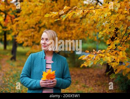 Schöne blonde Frau lächelt und schaut auf der linken Seite mit Ahornblättern in den Händen. Stockfoto