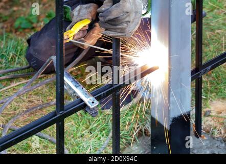 Helle Funken vom elektrischen Schweißen fliegen in verschiedene Richtungen gegen den Hintergrund in der Unschärfe des arbeitenden Schweißers und Gras. Stockfoto