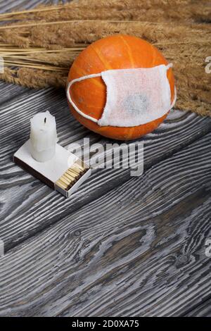 Orangefarbener Kürbis in einer medizinischen Maske. Liegt auf schwarzen Kiefernbrettern. In der Nähe gibt es einen Kerzenstumpf und ein Bouquet von getrocknetem Gras. Stockfoto