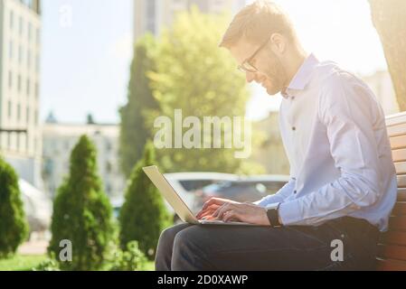 Seitenansicht eines schönen männlichen Freiberuflers oder Geschäftsmann arbeiten Online auf Laptop, während Sie auf der Bank im Freien sitzen Ein sonniger Tag Stockfoto