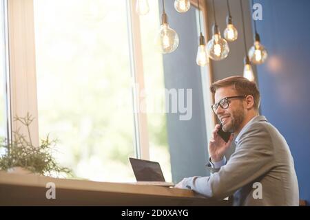Gute Nachrichten. Seitenansicht eines glücklichen jungen Geschäftsmannes, der per Telefon telefoniert und lächelt, während er im Café aus der Ferne arbeitet Stockfoto