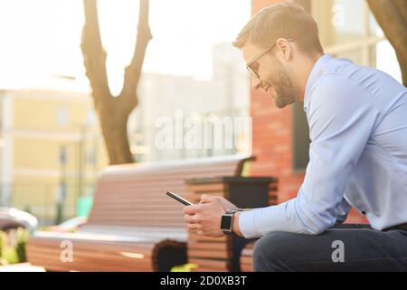 Chatten mit Freunden. Junger glücklicher Geschäftsmann in blauem Hemd und Brille mit Smartphone und lächelnd, während er auf der Bank auf einer sonnigen sitzen Stockfoto