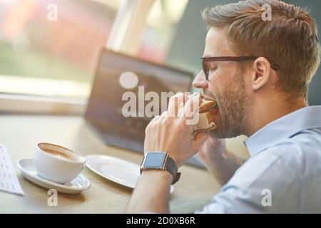 Seitenansicht eines jungen hungrigen Geschäftsmannes, der Sandwich isst Remote-Arbeit im Café Stockfoto