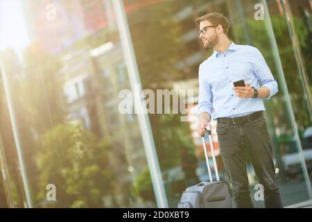 Warten auf Taxi. Eleganter, hübscher Geschäftsmann mit Koffer, der Smartphone hält und wegschaut, während er vor dem Flughafen steht Stockfoto
