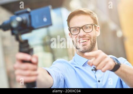 Junge glückliche männliche Blogger trägt blaues Hemd und Brillen mit einem Gimbal mit Smartphone halten, sprechen und lächeln an der Kamera während der Aufnahme Video für Stockfoto