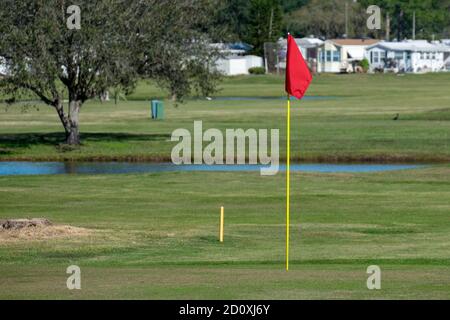 Eine rote Fahne steht bewegungslos an einem gelben Pol befestigt, an einem Golfloch, auf einem Golfplatz. Es gibt Wohnmobil Anhänger im Hintergrund. Stockfoto