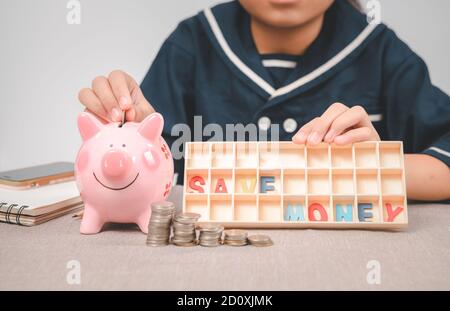 Kleines Mädchen setzen Münze in Sparschwein für das Sparen mit Haufen von Münzen auf dem Tisch zu Hause und zeigen Geld sparen Text. Ein rosa Sparschwein sind Lächeln und Hap Stockfoto