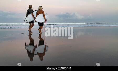 Zwei gutaussehende männliche Surfer, die am Strand spazieren, mit Surfbrettern, die sich bei Sonnenaufgang auf nassem Sand am Getreidestrand spiegeln. Bali, Indonesien Stockfoto