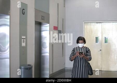 Geschäftsfrau trägt Gesichtsmaske Surfen ihr Handy, warten außerhalb Aufzug im Büro Gebäude Stockfoto