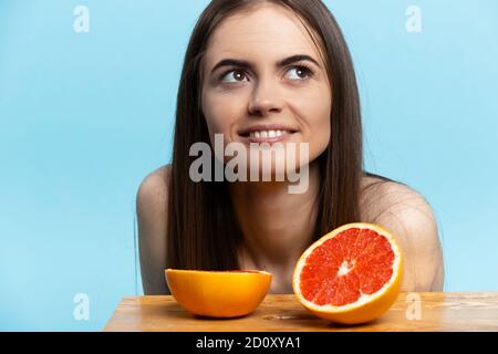 Lächelndes Mädchen, das nahe bei ihr aufschaut, ist zwei Hälften Grapefruit auf dem Holztisch. Blauer isolierter Hintergrund Stockfoto