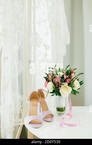 Lila Sandalen auf einem weißen Tisch am Fenster mit einem weißen Brautschleier und einem Blumenstrauß in einer Vase im Raum. Stockfoto